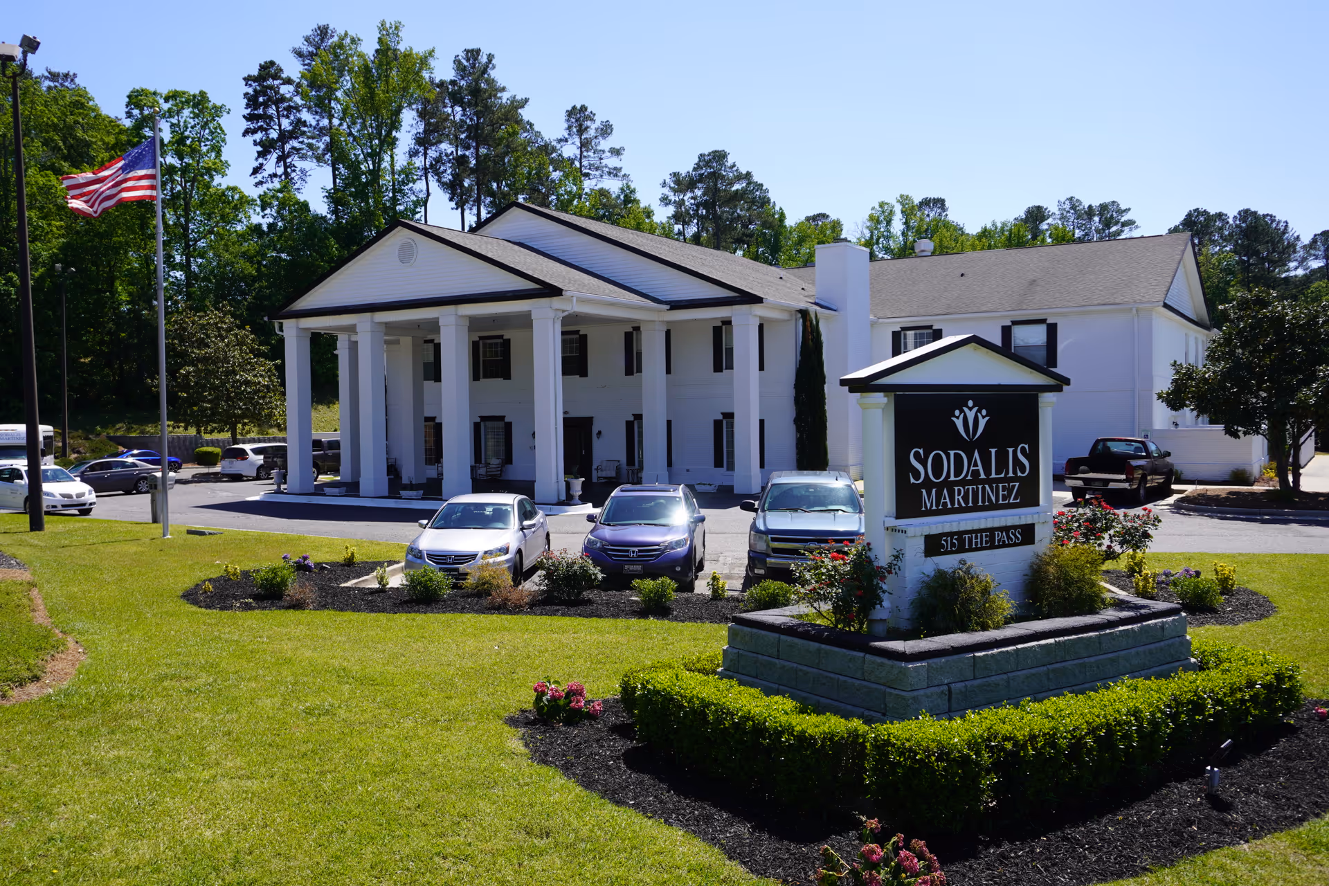 White two-story building with large columns and a front sign reading 'Sodalis Martinez' surrounded by a landscaped lawn and parked cars.