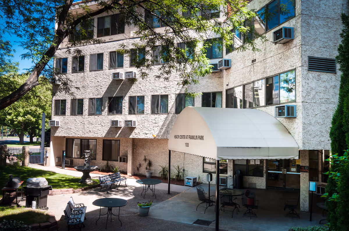Exterior view of the Health Center at Franklin Park building with a white canopy entrance, several windows with air conditioning units, outdoor seating with tables and chairs, a small fountain, and trees providing shade.