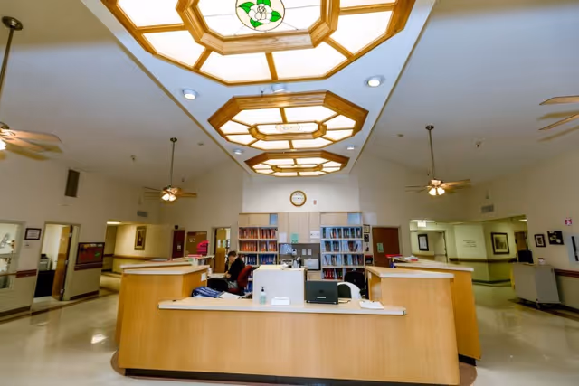 Reception area inside La Cañada Care Center with a large wooden desk, bookshelves filled with files, ceiling fans, and decorative ceiling lights. A staff member is seated behind the desk working on a computer.