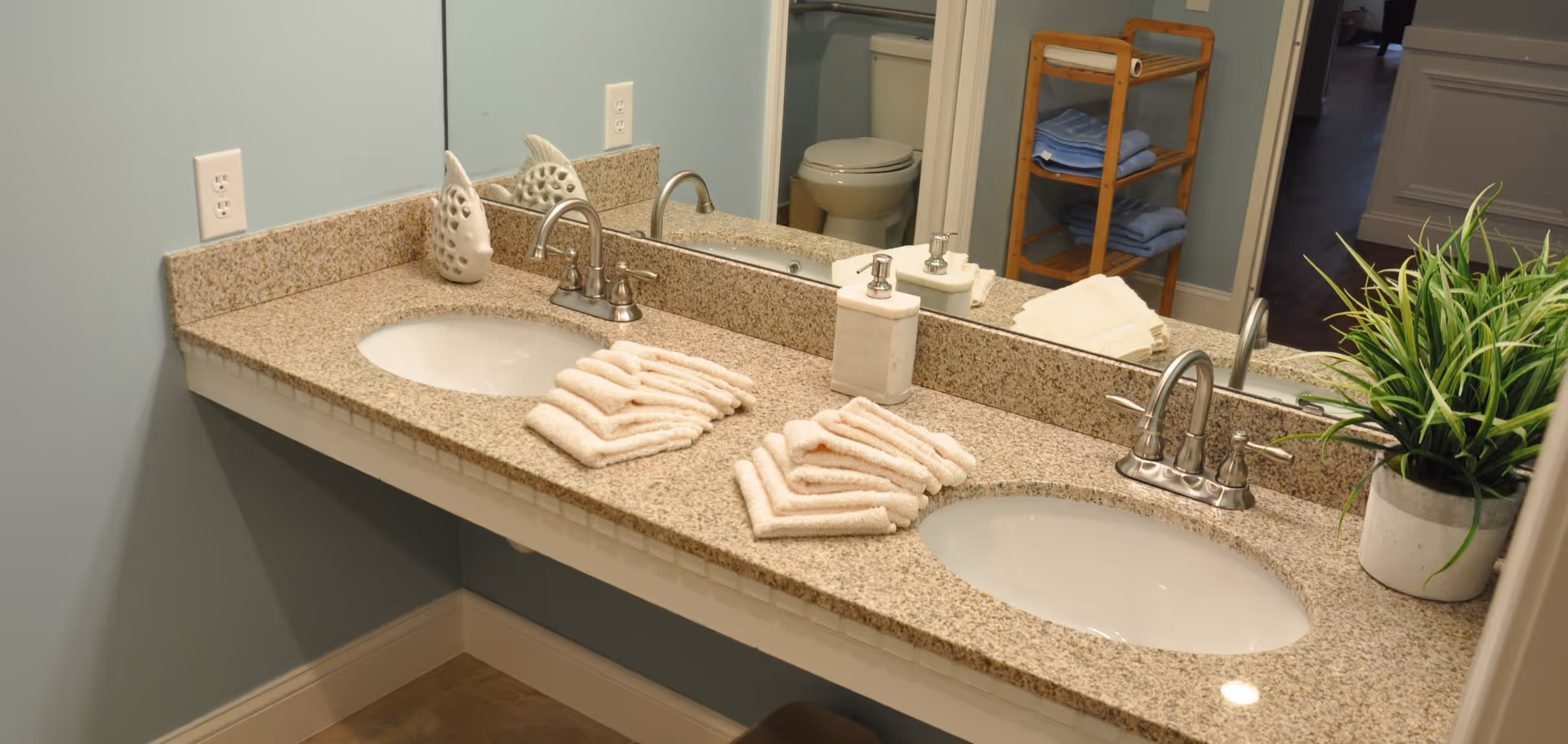 A bathroom countertop with two sinks and chrome faucets. On the countertop are neatly folded white towels, a soap dispenser, a decorative ceramic piece, and a potted green plant. A large mirror spans the length of the countertop, reflecting a toilet and a wooden shelf with folded towels in the background.