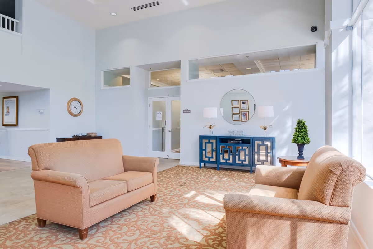 A bright and spacious sitting area in a senior living facility with two beige upholstered loveseats facing each other on a patterned carpet. A blue console table with decorative lamps and a round mirror is against the wall, next to a small round table with a potted plant. Large windows allow natural light to fill the room.