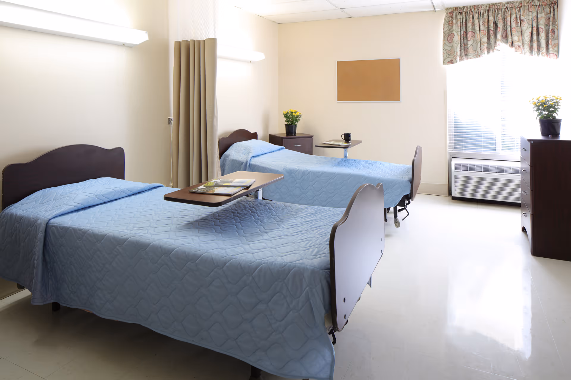 A bright and clean room with two single beds covered in light blue quilts. Each bed has a wooden headboard and a small overbed table. There are two dark wood nightstands with potted yellow flowers, a window with blinds and a floral valance, and a corkboard on the wall. The floor is shiny and light-colored, and the room is softly lit.