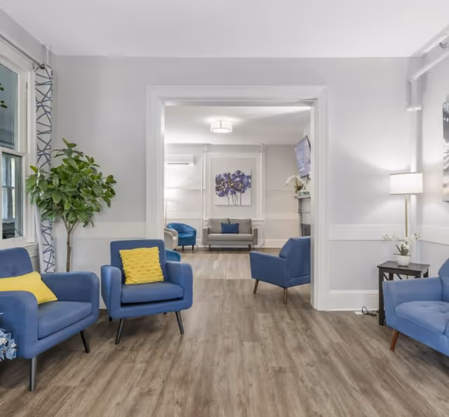 A bright and modern senior living community lounge area with blue armchairs featuring yellow cushions, a large green potted plant near a window with patterned curtains, light wood flooring, and white walls. Through a wide doorway, another seating area is visible with a gray sofa, blue armchairs, a wall-mounted TV, and a framed floral artwork.