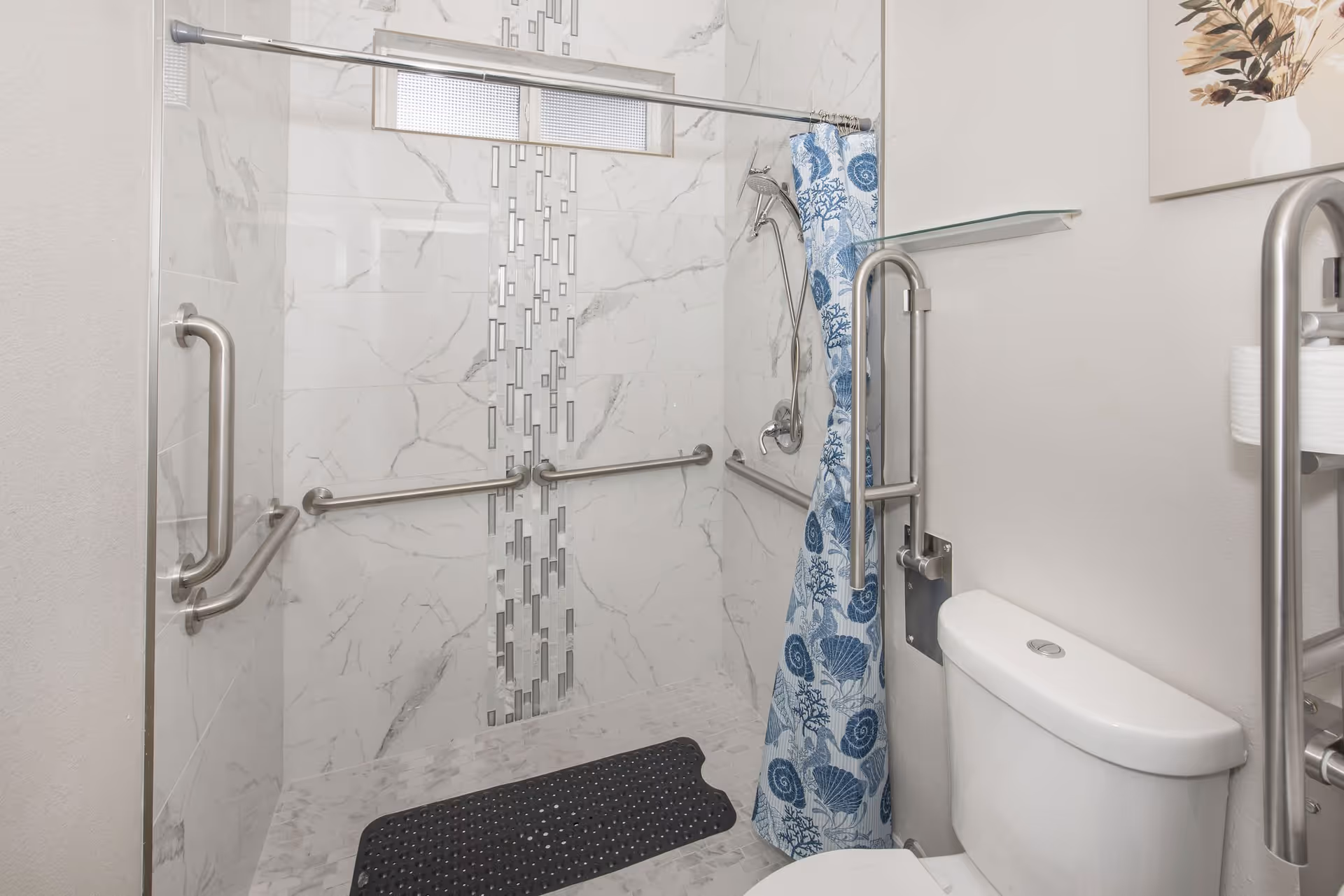 A bathroom with a walk-in shower featuring marble-patterned tiles and a decorative vertical strip of smaller tiles. The shower has multiple stainless steel grab bars and a handheld showerhead. A blue and white shower curtain with seashell and coral designs is partially drawn. A white toilet with a stainless steel grab bar next to it is visible, along with a small glass shelf mounted on the wall above the toilet. A framed artwork with dried flowers hangs on the wall.