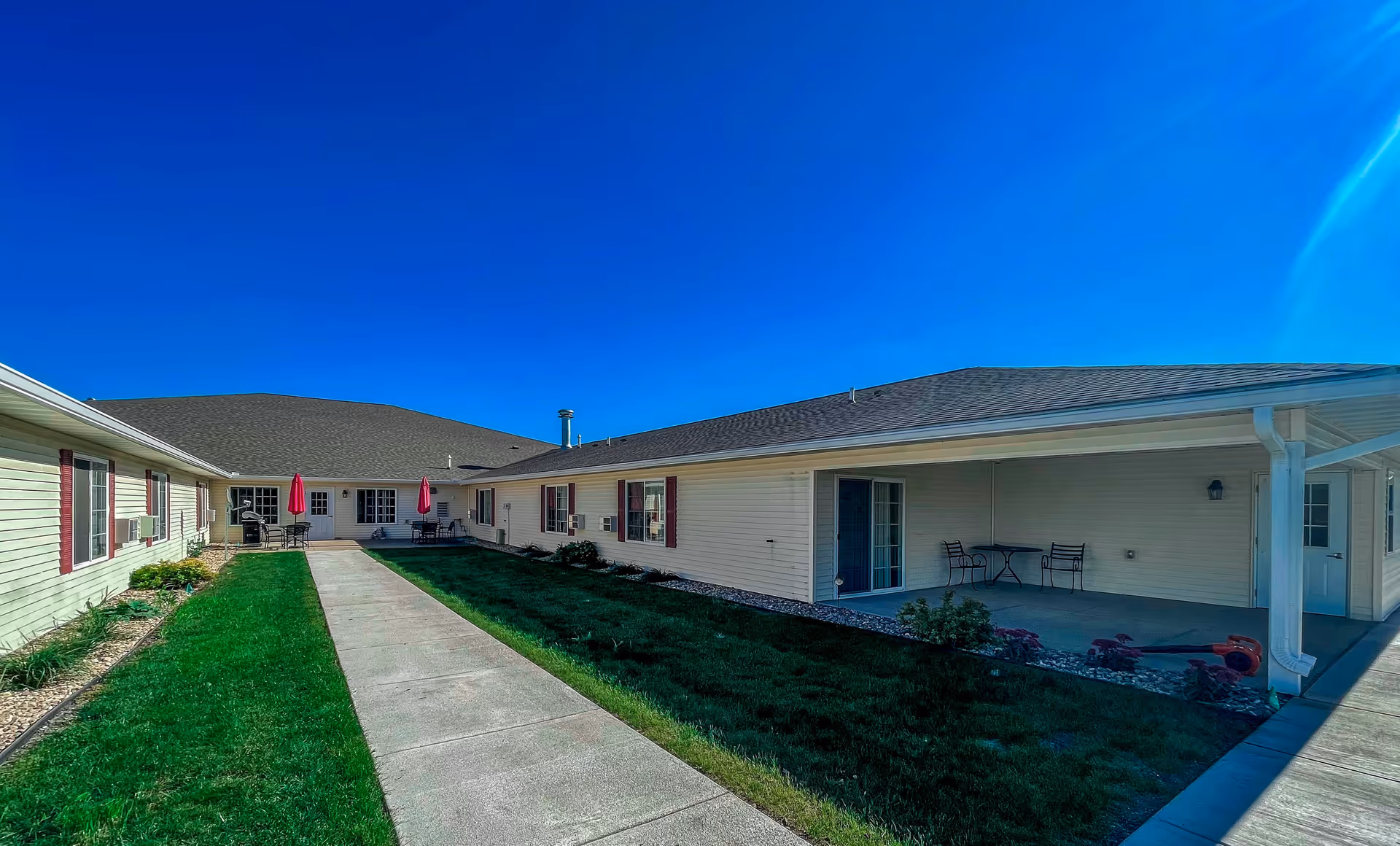 Outdoor courtyard area of a senior living facility with a concrete walkway, green grass on both sides, and beige buildings with red shutters. There are patio tables with red umbrellas and chairs near the building walls under a clear blue sky.