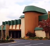 Exterior view of a building with a unique architectural design featuring a curved green roof and tan walls, surrounded by landscaped bushes and a tree with red leaves, under a partly cloudy sky.