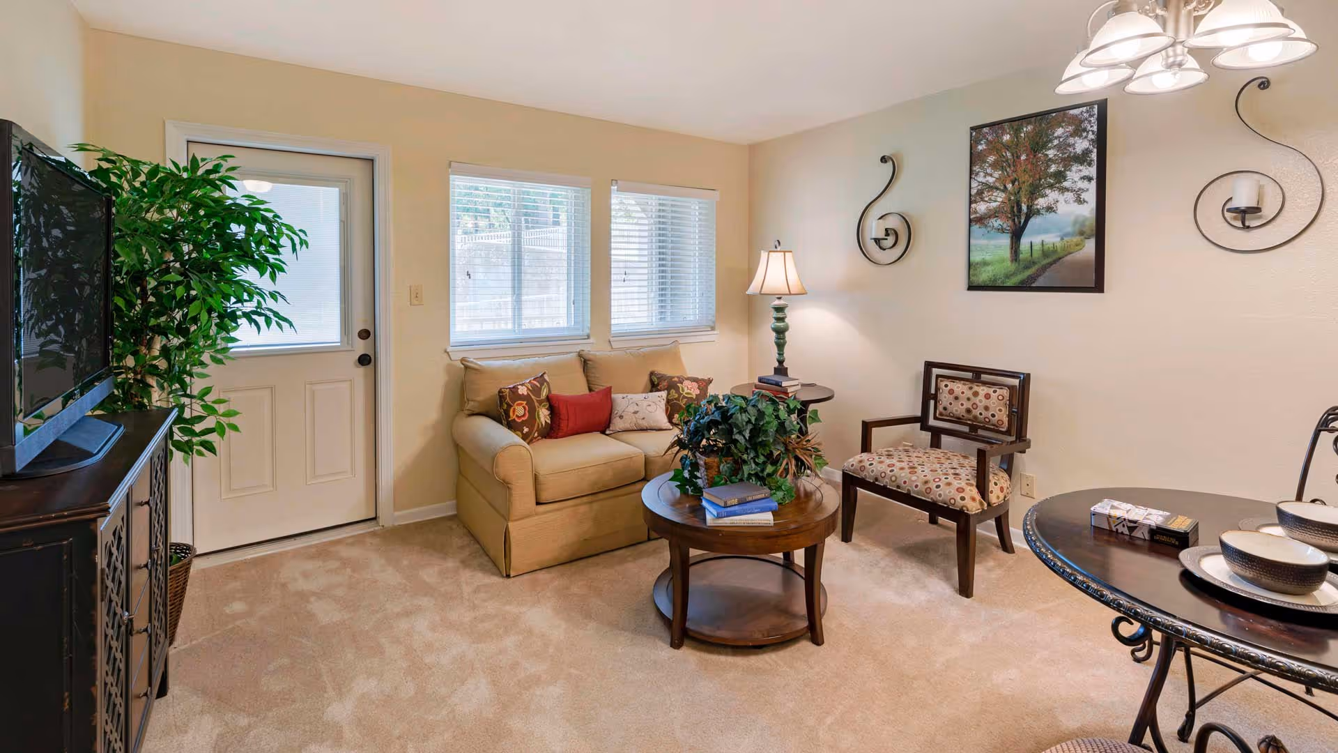 A cozy living room with beige walls and carpet, featuring a beige loveseat with colorful pillows, a wooden round coffee table with a plant and books, a patterned armchair, a side table with a lamp, a wall painting of a tree, and a TV on a dark wooden stand next to a door and two windows with blinds.