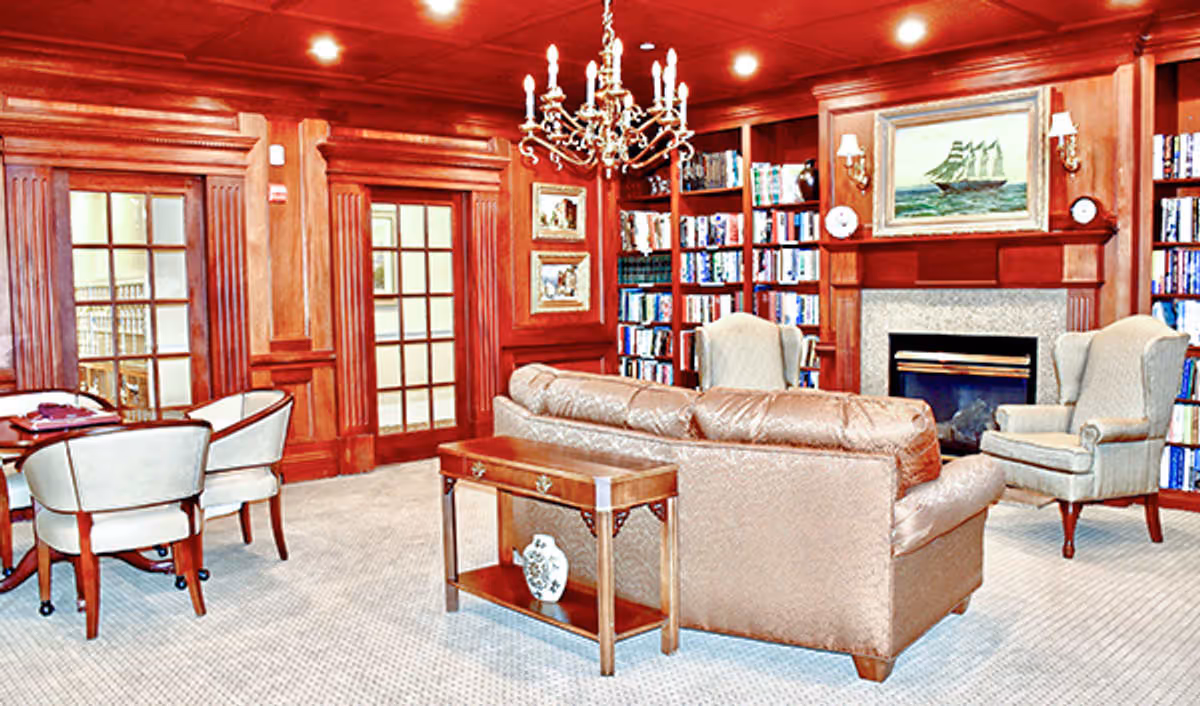 A cozy living room with wood-paneled walls and built-in bookshelves filled with books. The room features a leather sofa, two upholstered armchairs, a wooden side table with a decorative vase, and a fireplace with a painting of a sailing ship above it. A chandelier hangs from the ceiling, and there is a small round table with four chairs near glass-paneled double doors.