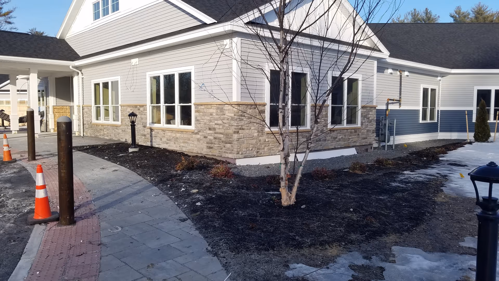 Exterior view of a building with light gray siding and stone accents, a paved walkway with orange traffic cones, a small tree with no leaves, and some landscaping with mulch and small shrubs. The sky is clear and blue.