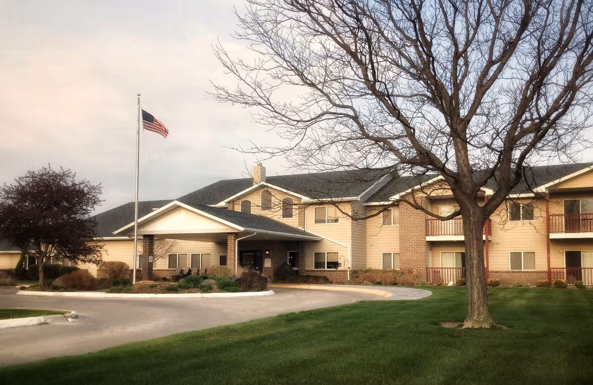 Front exterior of the Ridgeway Senior Living building with a covered entrance, American flag on a flagpole, driveway and a large tree on the lawn.