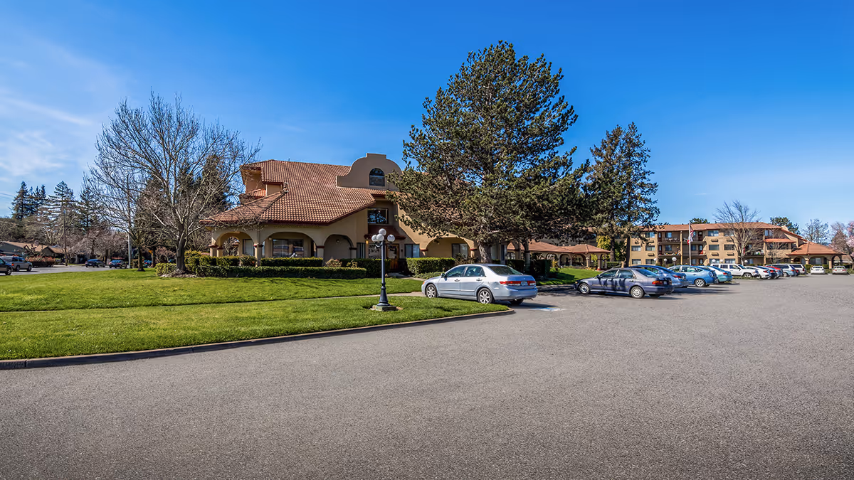 Parking lot and green lawn in front of a multi-story Mediterranean-style senior living building with cars and trees under a blue sky.