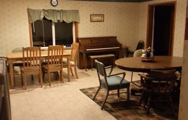 Dimly lit senior living dining area with tables, chairs, piano, and wallpapered walls.