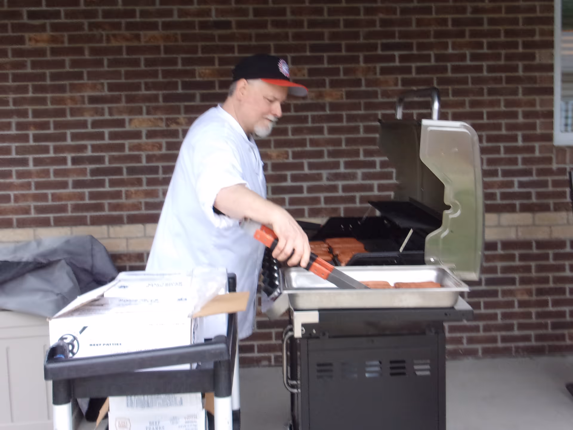 A man wearing a white shirt and a black and red cap is grilling hot dogs on an outdoor grill against a brick wall background. There is a cart with boxes next to him.