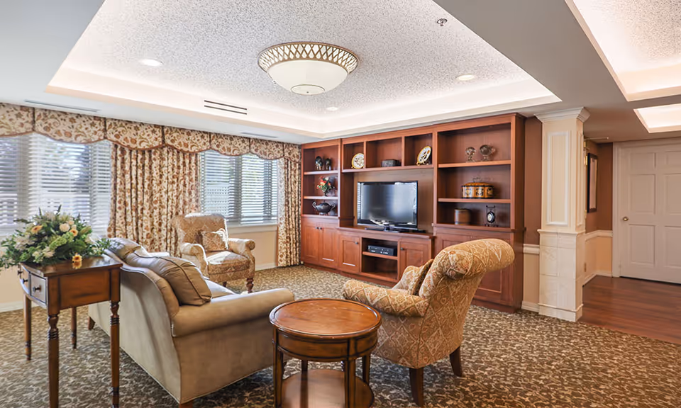Cozy living room with upholstered sofa and armchairs, a wooden entertainment center with a TV, patterned carpet and draped windows.
