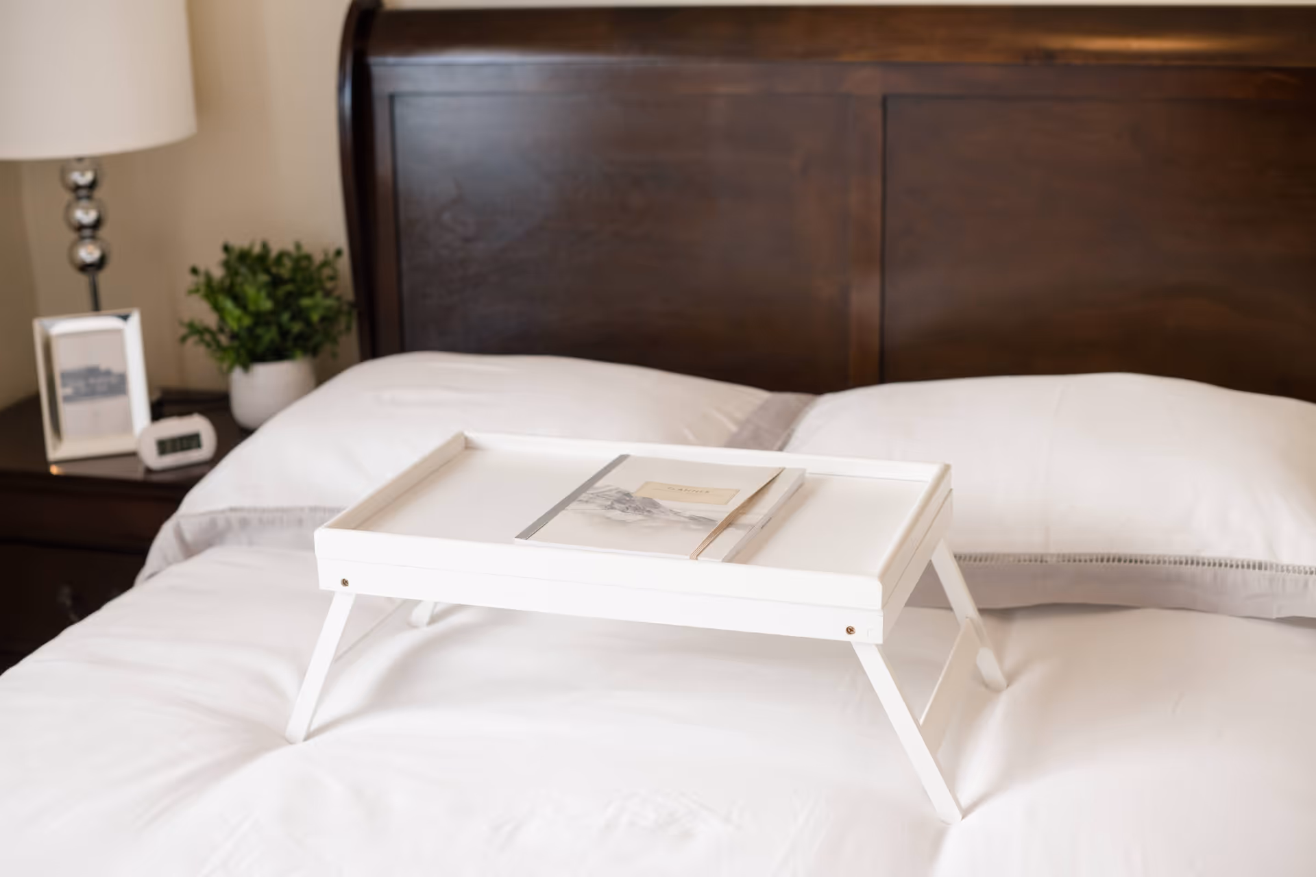A neatly made bed with white linens and a dark wooden headboard. On the bed is a white foldable tray table holding a closed notebook. To the left of the bed is a nightstand with a lamp, a small green plant, a framed photo, and a digital clock.