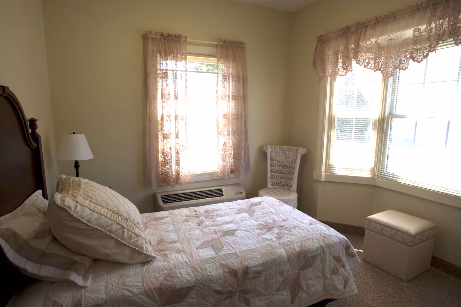 Sunlit bedroom with a single bed covered in a pastel quilt, lace-curtained windows, a wicker chair, floor lamp, and an upholstered ottoman.
