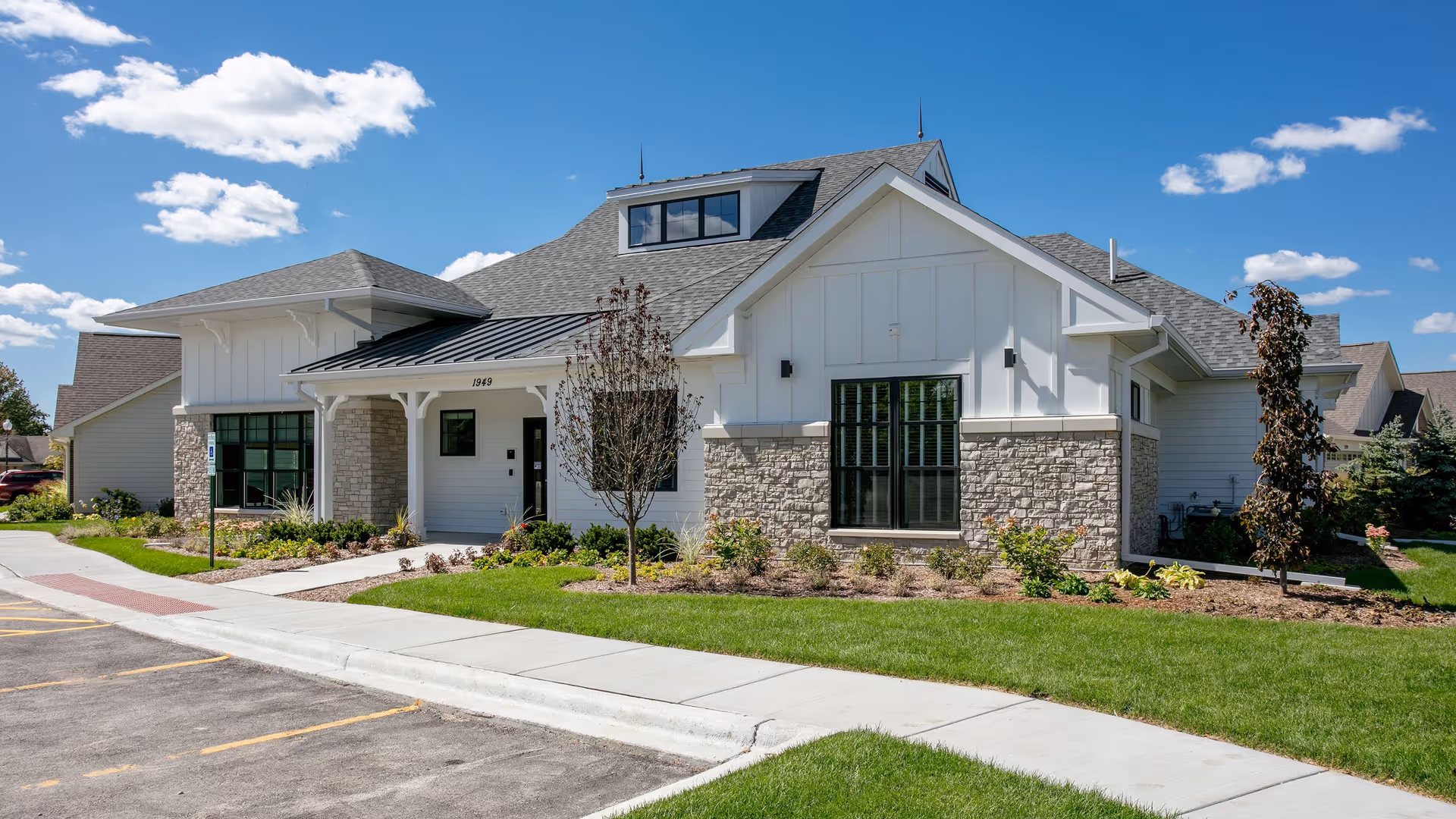 Exterior view of a single-story building with a combination of white siding and stone facade, surrounded by a well-maintained lawn and small trees under a blue sky with scattered clouds.