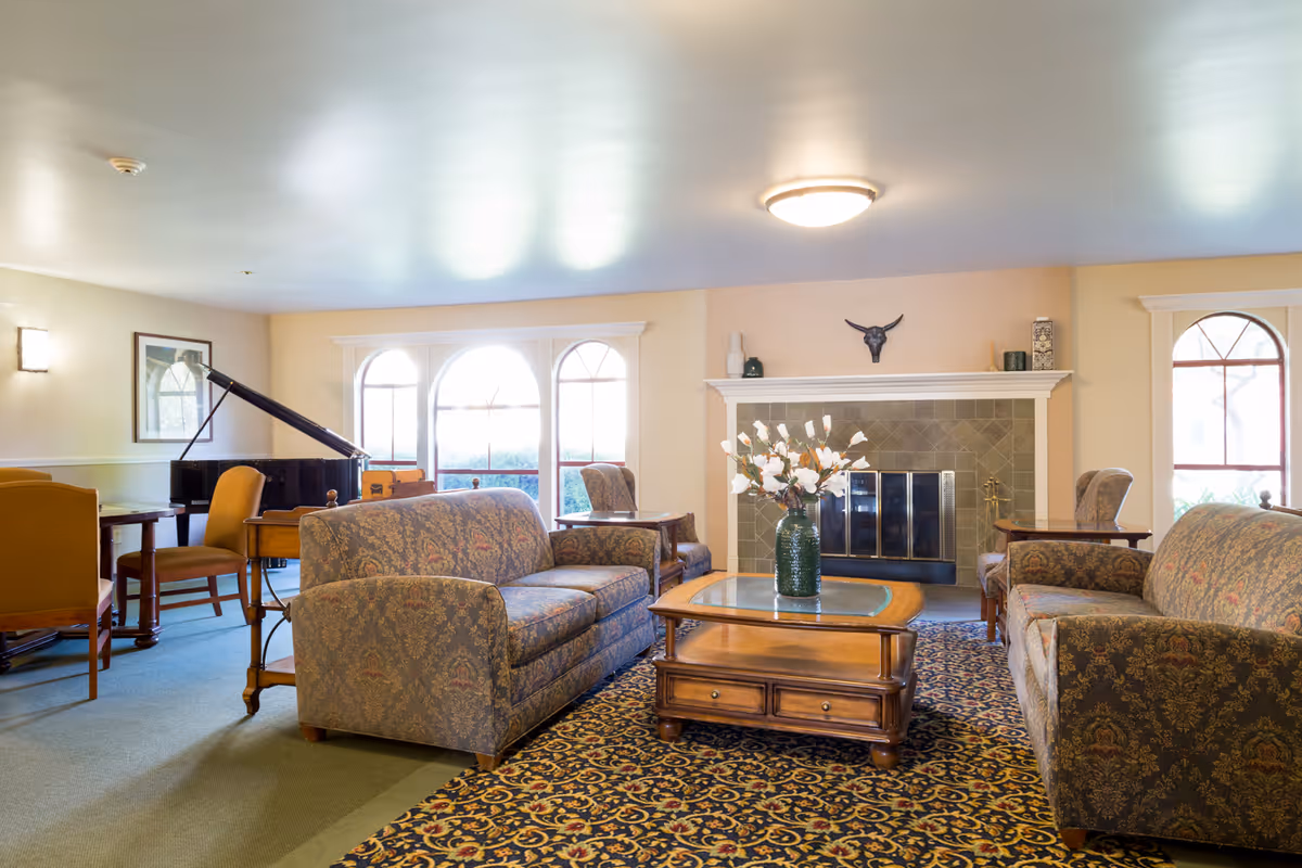 A cozy living room area in a senior living facility with two patterned sofas facing each other across a wooden coffee table with a vase of flowers. The room features a fireplace with decorative items on the mantel, large arched windows letting in natural light, a patterned carpet, and a grand piano in the background.