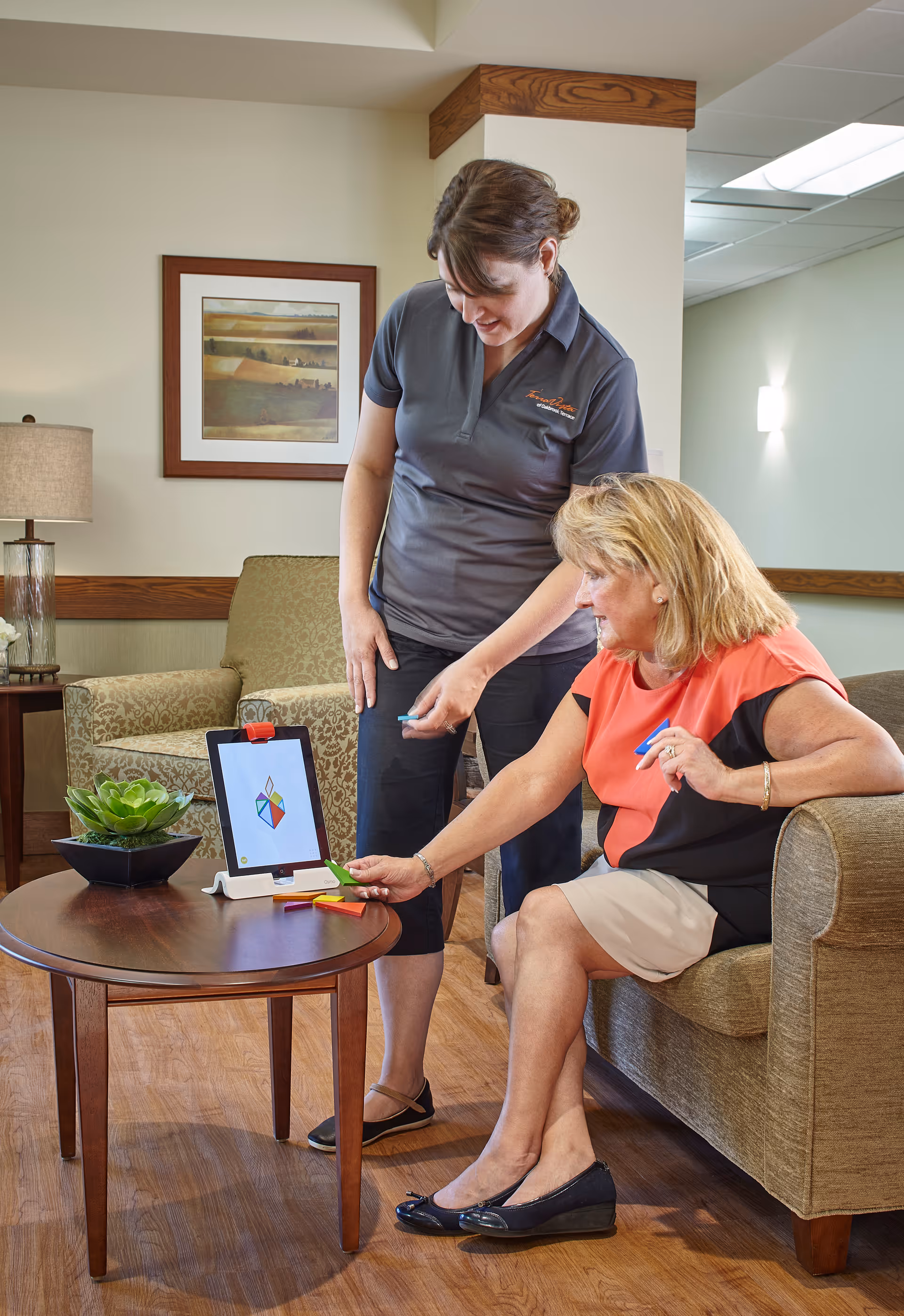 A woman sitting on a couch interacts with a tablet on a wooden coffee table while another woman stands beside her, both engaged in an activity in a cozy living room setting with a framed picture on the wall, a lamp, and an armchair in the background.
