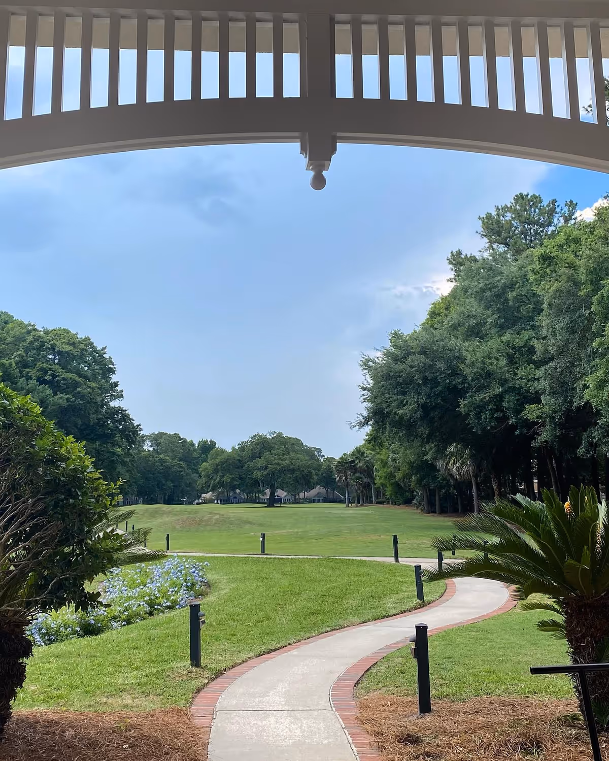 View of a winding concrete pathway bordered with red bricks leading through a well-maintained grassy area with trees and shrubs on either side, seen from under a white pergola or covered structure. The sky is partly cloudy.