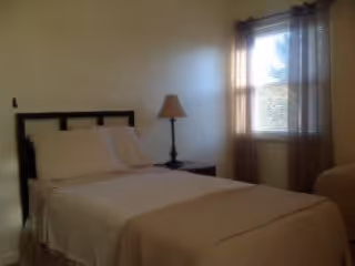 A simple bedroom with a neatly made bed featuring white bedding and pillows, a dark wooden headboard, a bedside table with a lamp, and a window with sheer curtains allowing natural light to enter.