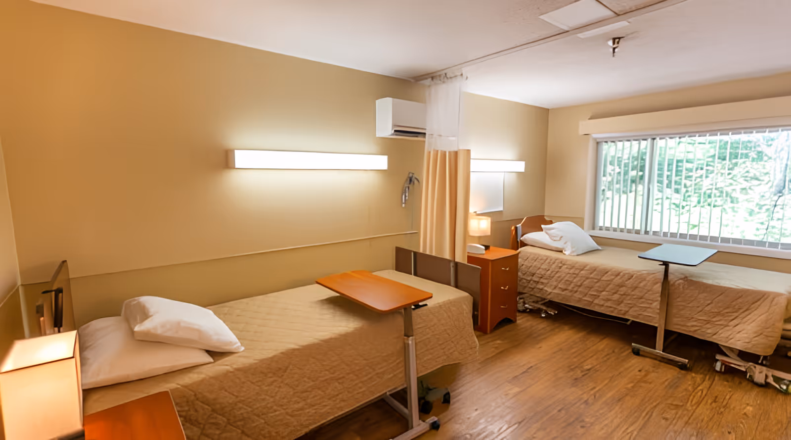 A room with two single beds, each with beige bedspreads and white pillows. Each bed has a small overbed table on wheels. There is a wooden nightstand with a lamp between the beds. The room has beige walls, a large window with vertical blinds, and wooden flooring. A curtain hangs from the ceiling between the beds for privacy.