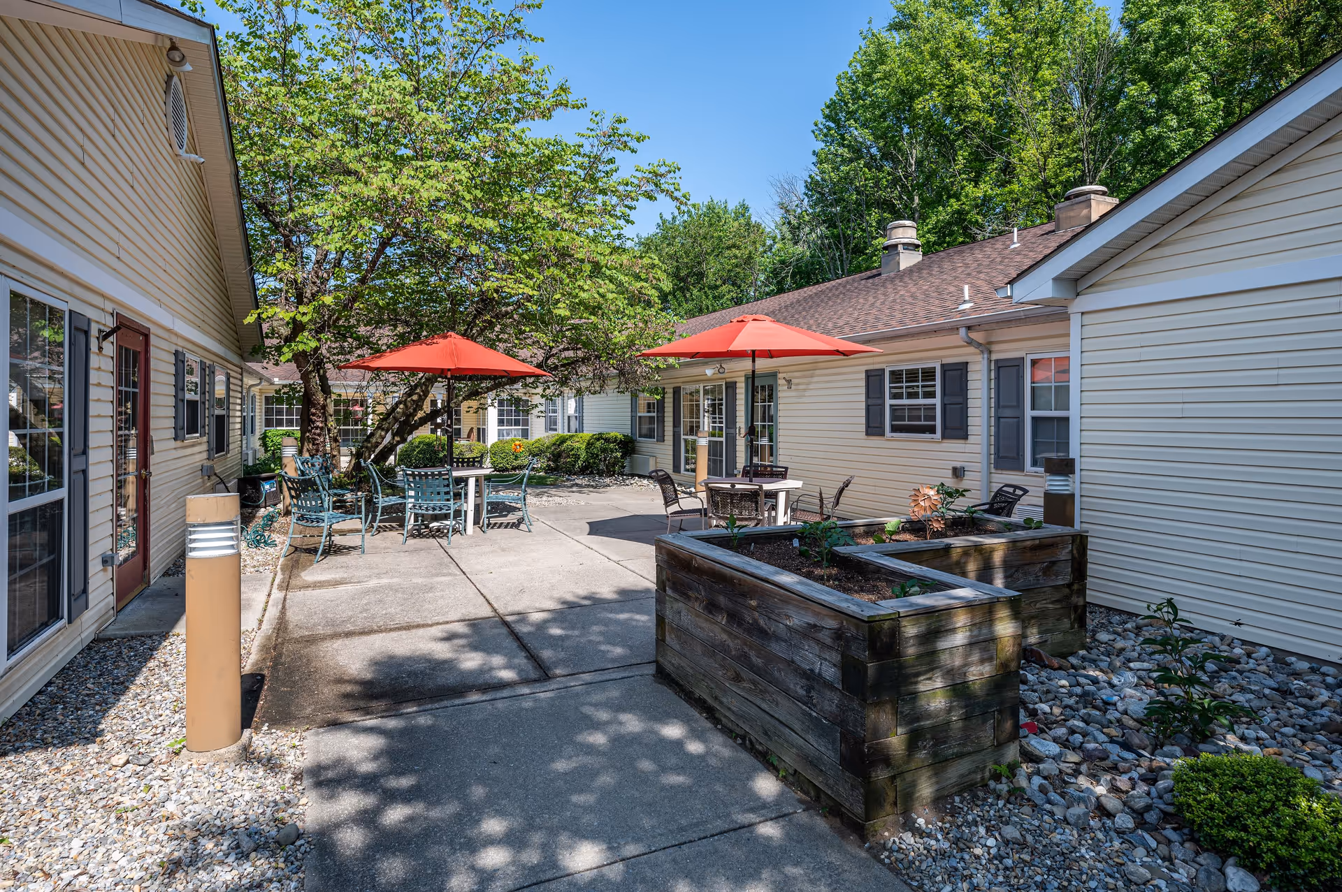 Outdoor courtyard area of a senior living facility with beige siding buildings surrounding a concrete patio. The patio has several metal tables and chairs, some shaded by red umbrellas. There are raised wooden garden beds with plants and trees providing shade. The sky is clear and blue.