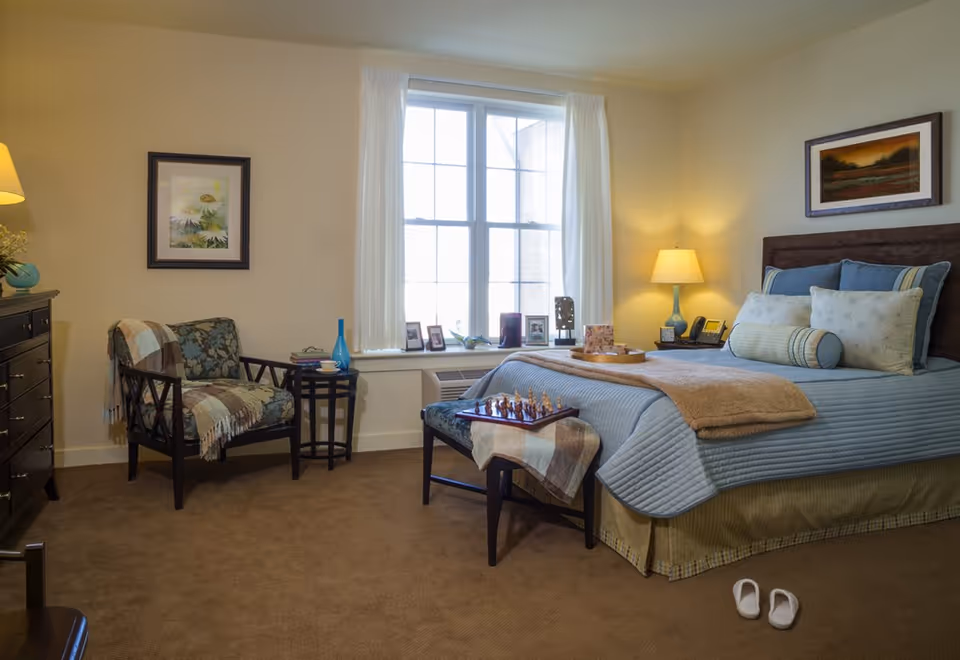 A cozy bedroom with a neatly made bed featuring blue and beige bedding, a small bench at the foot of the bed with a chessboard on it, a cushioned armchair with a throw blanket, a side table with a blue vase, a dresser, framed artwork on the walls, and a window with white curtains letting in natural light. A pair of white slippers is placed on the carpeted floor near the bed.
