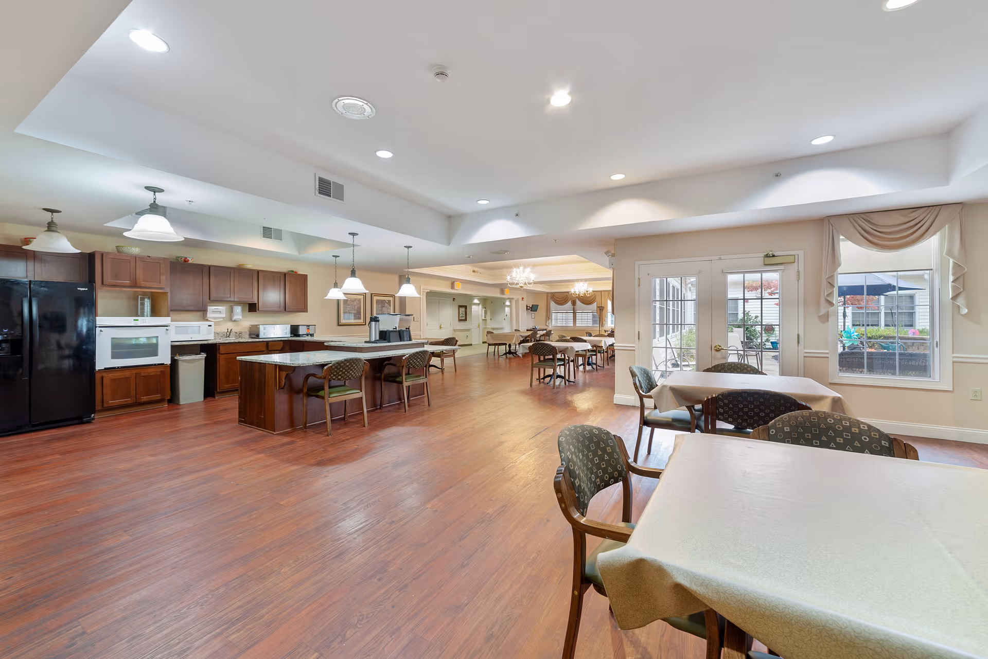 Spacious dining area with multiple tables and chairs, adjacent to a kitchen with wooden cabinets, black refrigerator, white oven, and microwave. The room has wooden flooring, ceiling lights, and large windows with drapes letting in natural light. A door leads to an outdoor patio area with seating and umbrellas.