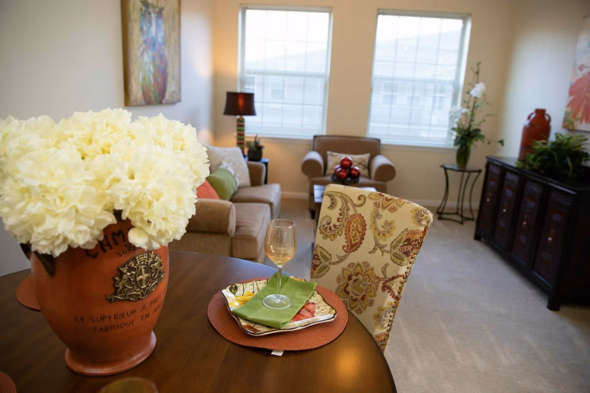 A cozy living room area with a round wooden table in the foreground set with a decorative plate, green napkin, and a wine glass. A large terracotta vase with white flowers sits on the table. In the background, there is a beige sofa with colorful cushions, a brown armchair, a side table with a lamp, and a dark wooden cabinet with plants and decorative items. Two large windows let in natural light.