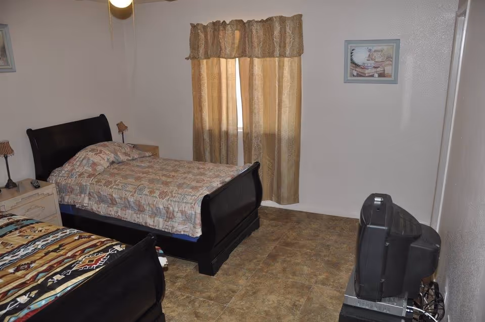 A bedroom with two single beds featuring dark wooden frames and patterned bedspreads. There are two bedside tables with lamps, a window with beige curtains, a framed picture on the wall, and an older-style CRT television on a stand in the corner.
