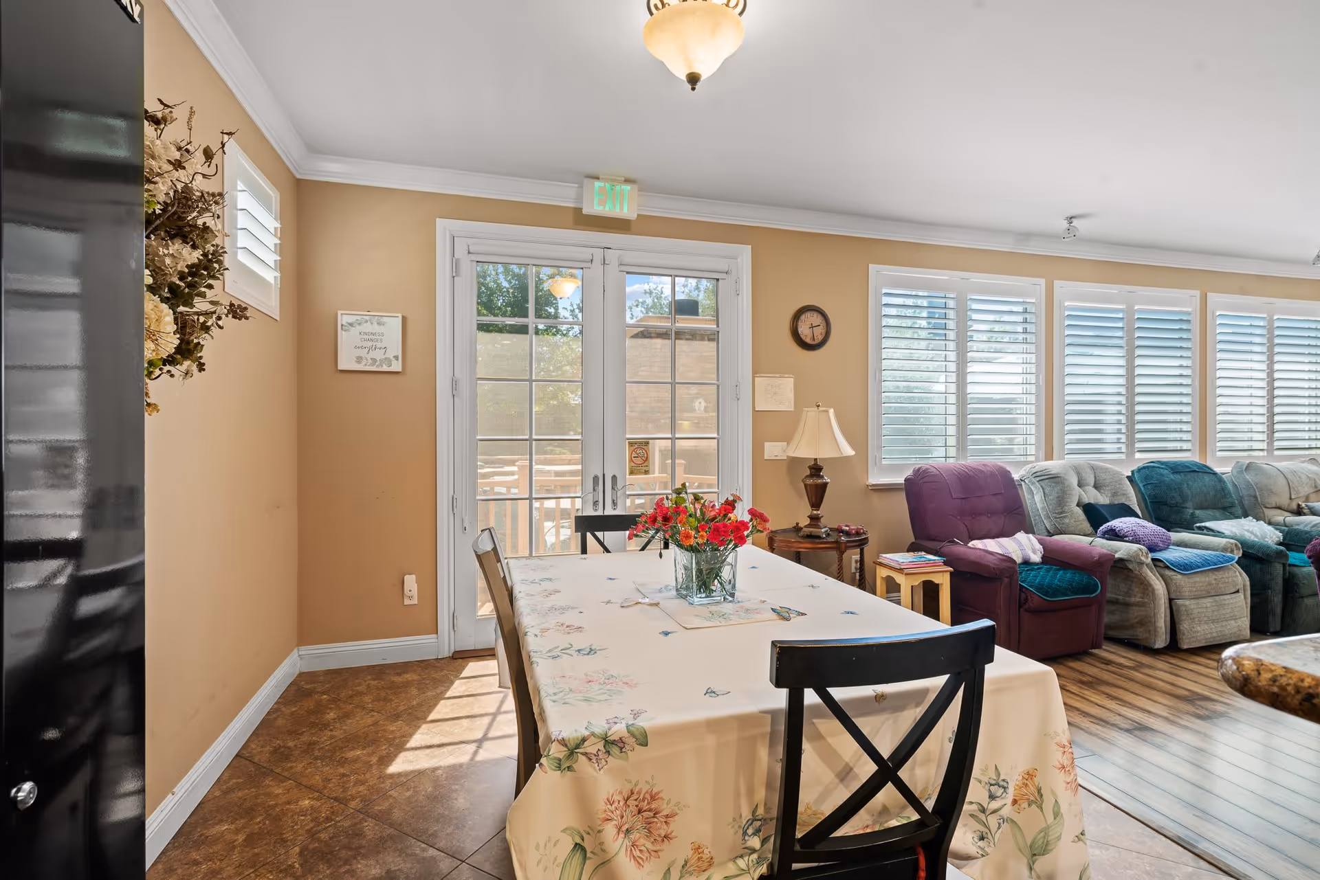 A bright and cozy living and dining area with a table covered in a floral tablecloth and a vase of red flowers in the center. Behind the table are large windows with white shutters and a row of comfortable recliner chairs in various colors. A lamp and small side table are positioned near the chairs, and a glass door leads outside, letting in natural light.