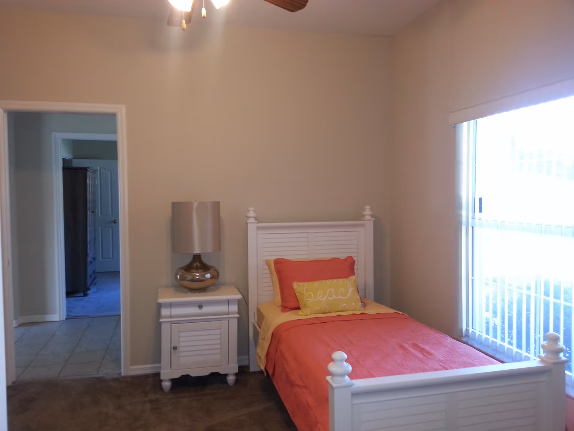 A small bedroom with a white wooden single bed featuring coral and yellow bedding and pillows. Next to the bed is a white nightstand with a metallic lamp. There is a large window with vertical blinds on the right side, and an open doorway leading to another room on the left.