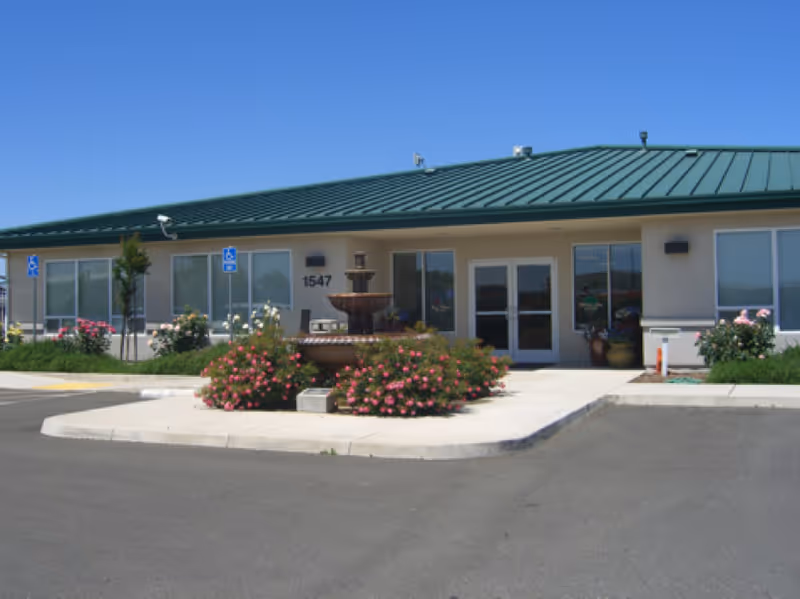 Single-story care center building with a green metal roof, central fountain and flower beds in front of the main entrance numbered 1547 under a clear blue sky.