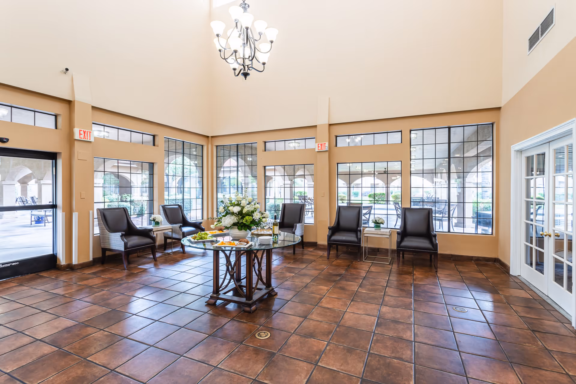 A spacious, well-lit common area with large windows and glass doors showing an outdoor patio. The room features a round glass table with a floral centerpiece and snacks, surrounded by six dark leather chairs. The floor is tiled with brown tiles, and a chandelier hangs from the high ceiling.