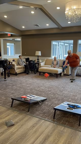 A group of elderly people in a spacious, well-lit living room with beige walls and carpeted floor. Some are seated on cream-colored sofas and chairs, with wheelchairs nearby. One elderly man is standing and playing a game of cornhole, tossing a bean bag towards a cornhole board on the floor. The room has large windows, a chandelier, and recessed ceiling lights.