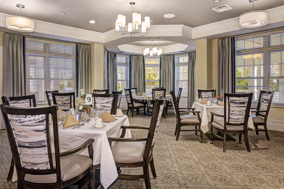 A bright and elegant dining room with multiple tables set with white tablecloths, beige napkins, cups, glasses, and silverware. The room features large windows with blinds and curtains, patterned carpet, and modern chandeliers hanging from the ceiling.
