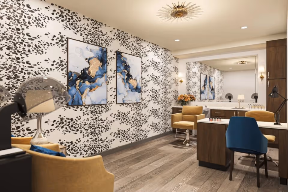 A stylish interior room with patterned wallpaper featuring black leaf-like designs on a white background. Two abstract blue and gold paintings hang on the wall. The room has wooden flooring and modern furniture including a yellow armchair, a blue chair, and a wooden desk with a lamp. A large mirror reflects the room, and there is a hair dryer hood visible on the left side.
