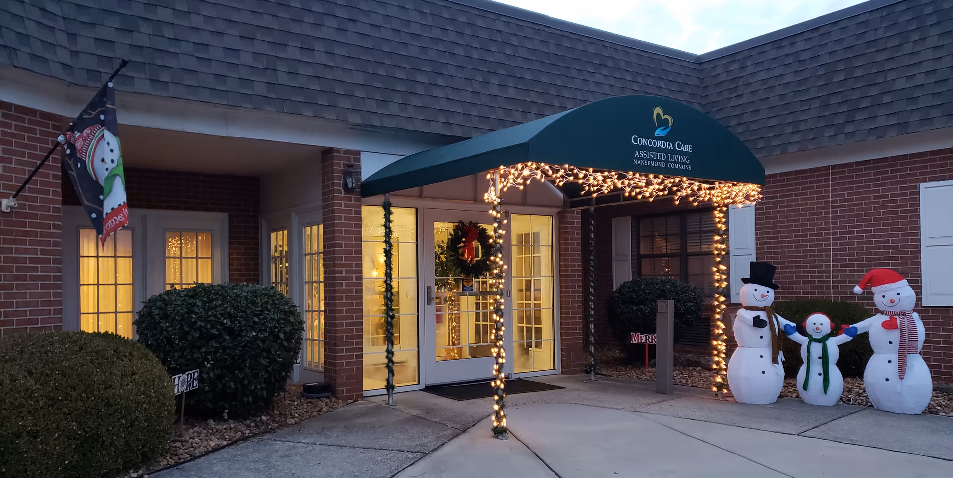 Entrance of Concordia Care Assisted Living Nansemond Commons decorated for the holidays with a green awning, string lights, a wreath on the door, a snowman family decoration, and a festive flag on the left side.
