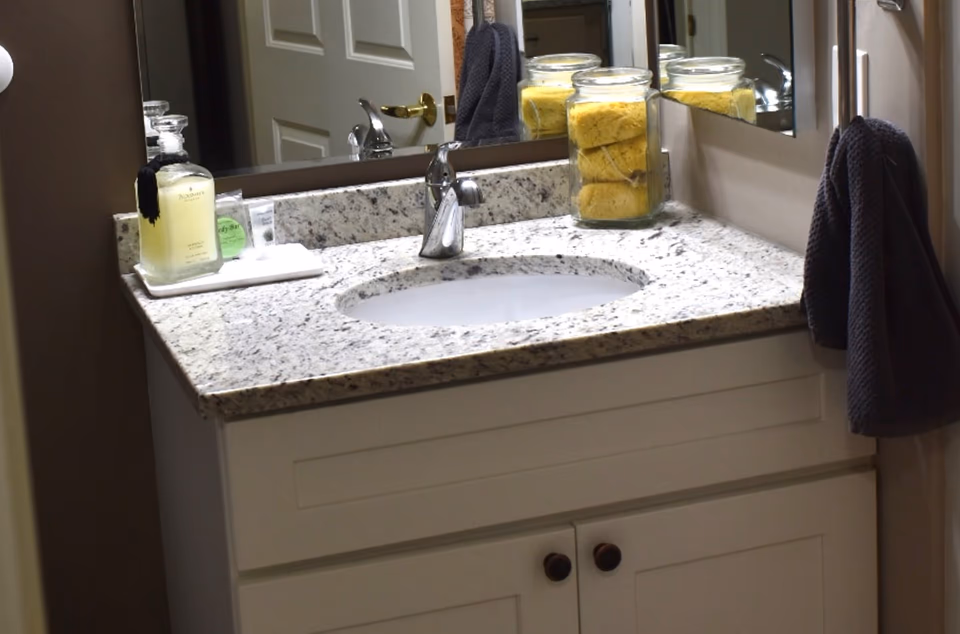 Bathroom vanity with a granite countertop, an oval sink, a chrome faucet, a glass jar filled with yellow sponges, a soap dispenser, a small tray with toiletries, a dark towel hanging on a hook, and a mirror reflecting a door with a gold handle.