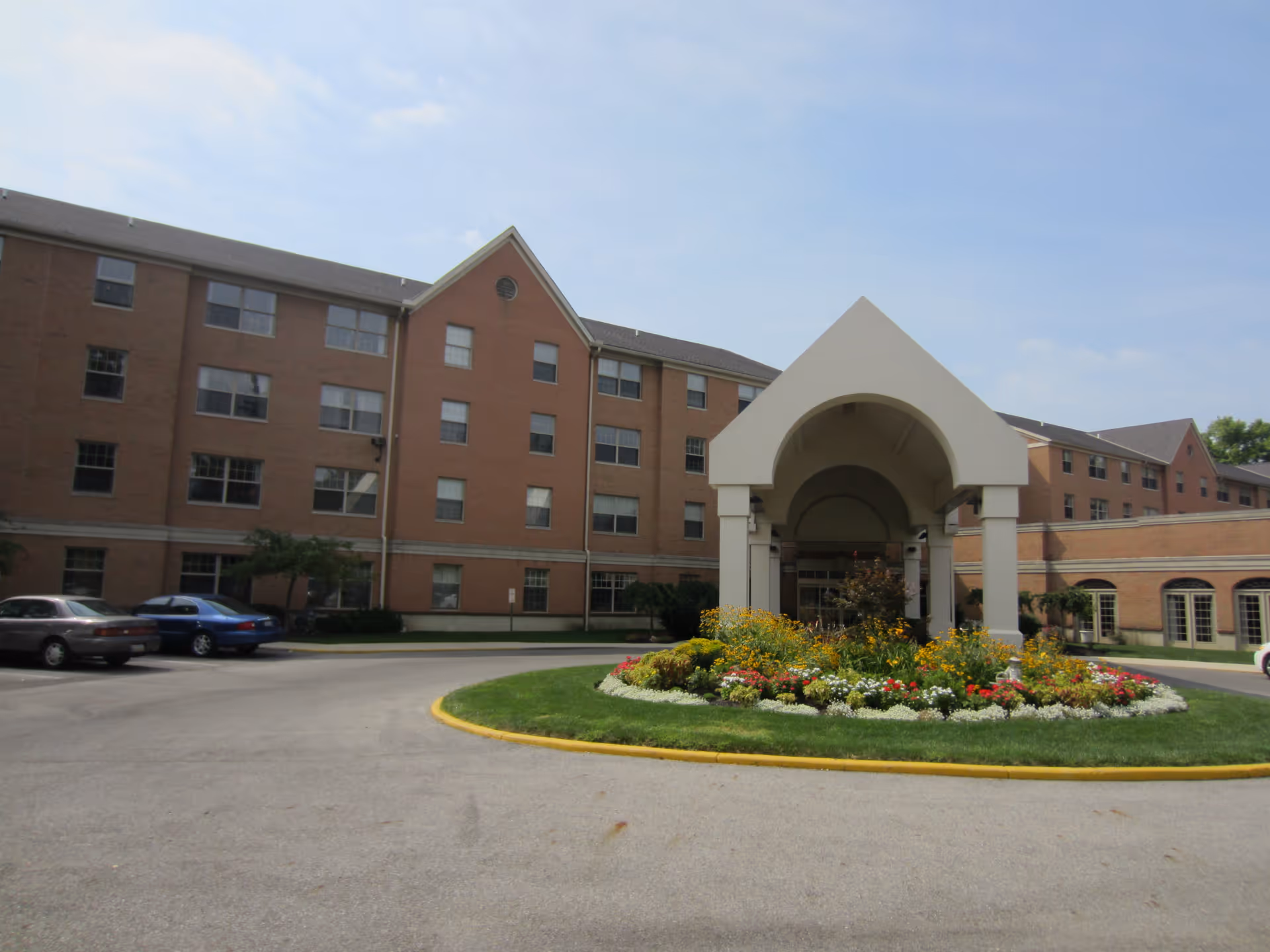 Exterior view of a multi-story brick building with a covered entrance and a circular flower bed in front. Several cars are parked along the driveway, and the sky is partly cloudy.