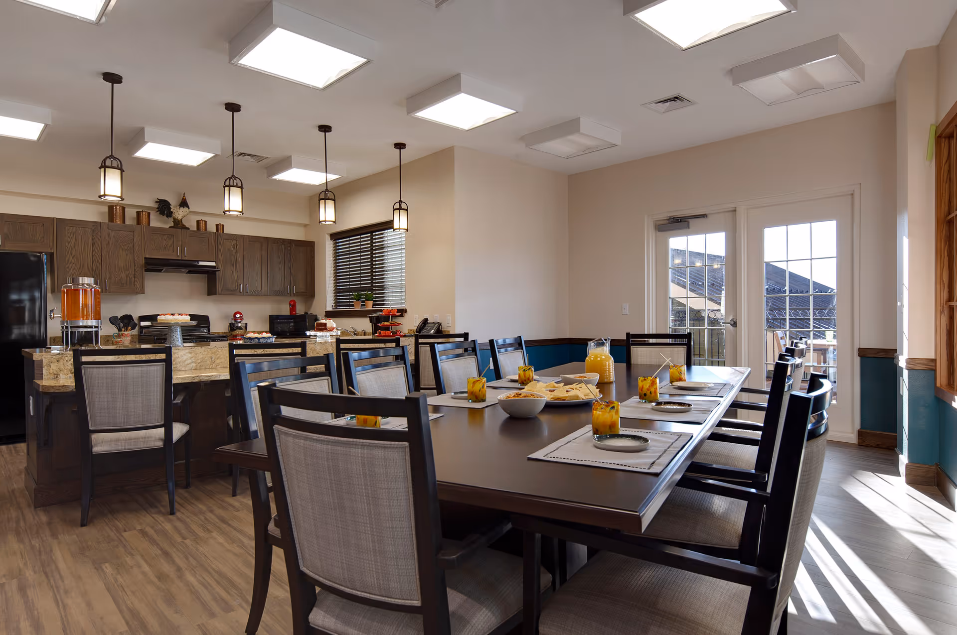 Well-lit dining area with a long table set with plates and glasses, adjacent to an open kitchen with pendant lights and cabinetry.