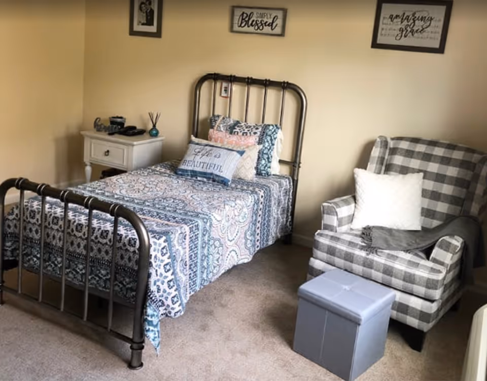 A small bedroom with a metal-framed twin bed covered in a patterned quilt, a white nightstand, and a gray plaid armchair with ottoman.
