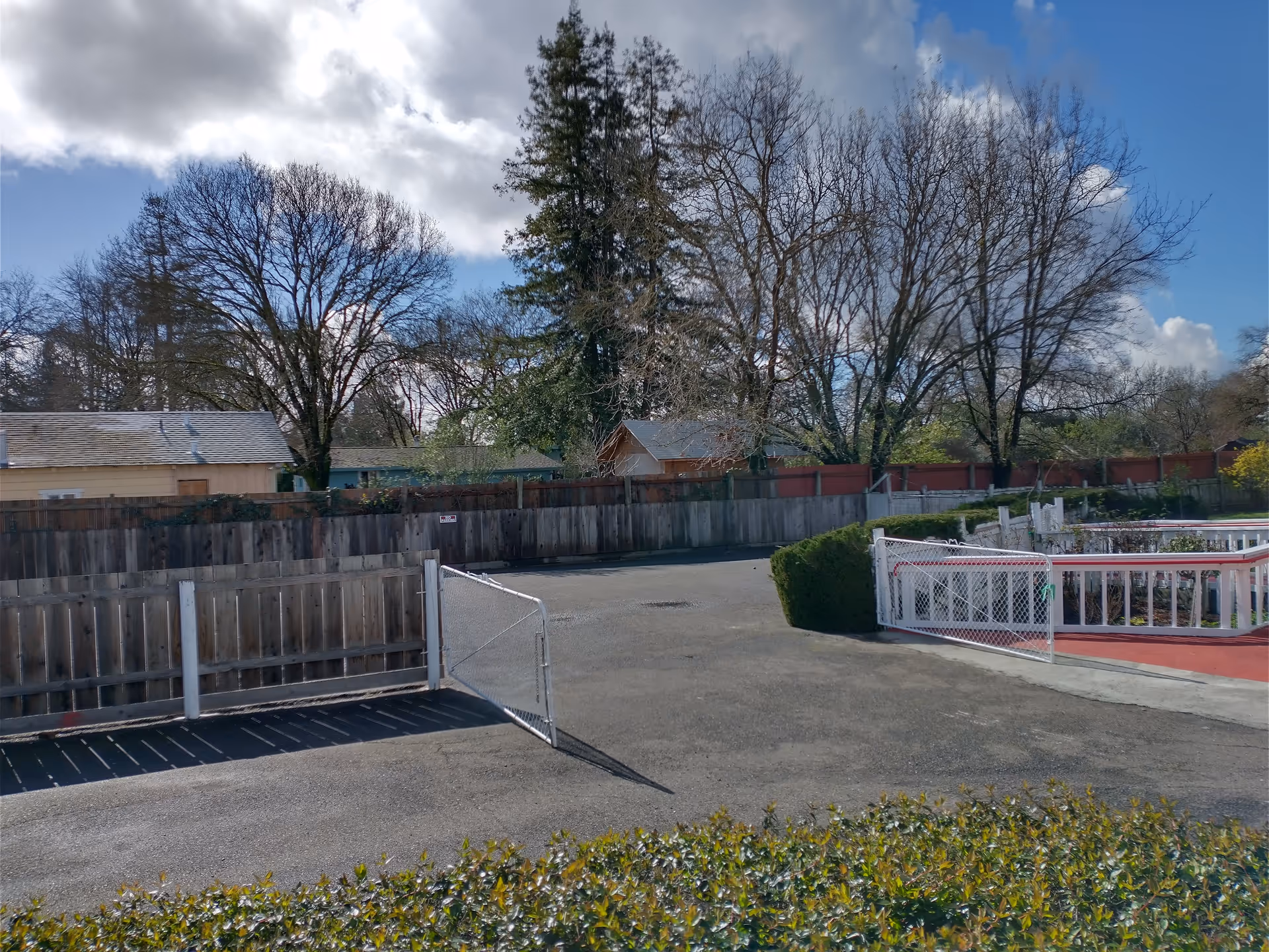 Outdoor view of a fenced area with a partially open metal gate, surrounded by wooden fences and leafless trees under a partly cloudy sky.
