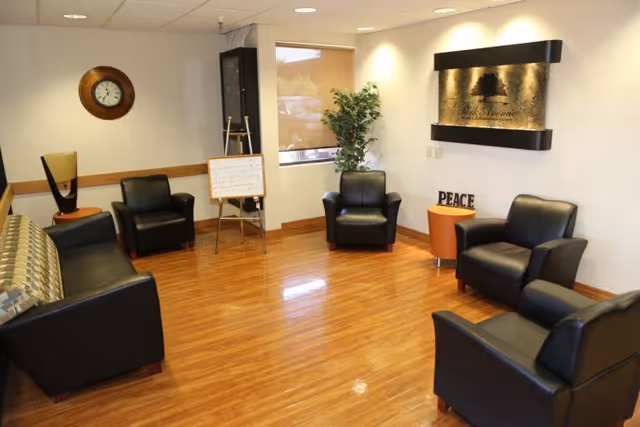 A waiting or lounge area with wooden flooring, black leather chairs and a sofa arranged around the room. There is a wall clock, a potted plant, a small round orange table with the word 'PEACE' on it, and a sign on the wall that reads 'Park Avenue Health And Rehabilitation'.