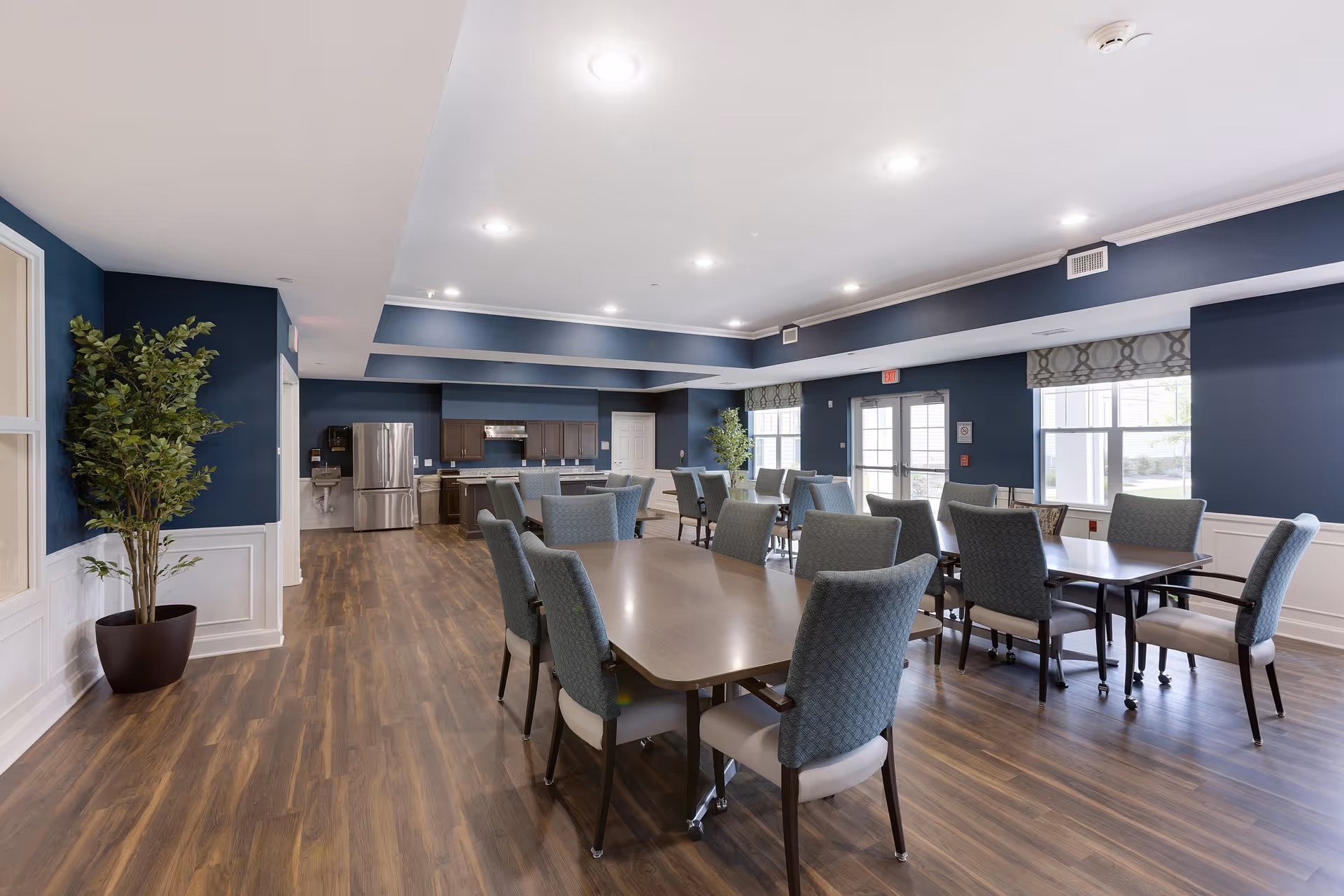 A spacious dining room with several tables and chairs arranged neatly. The room features wood flooring, blue walls with white wainscoting, and large windows allowing natural light. In the background, there is a kitchen area with stainless steel appliances including a refrigerator and a stove. Potted plants are placed near the walls for decoration.