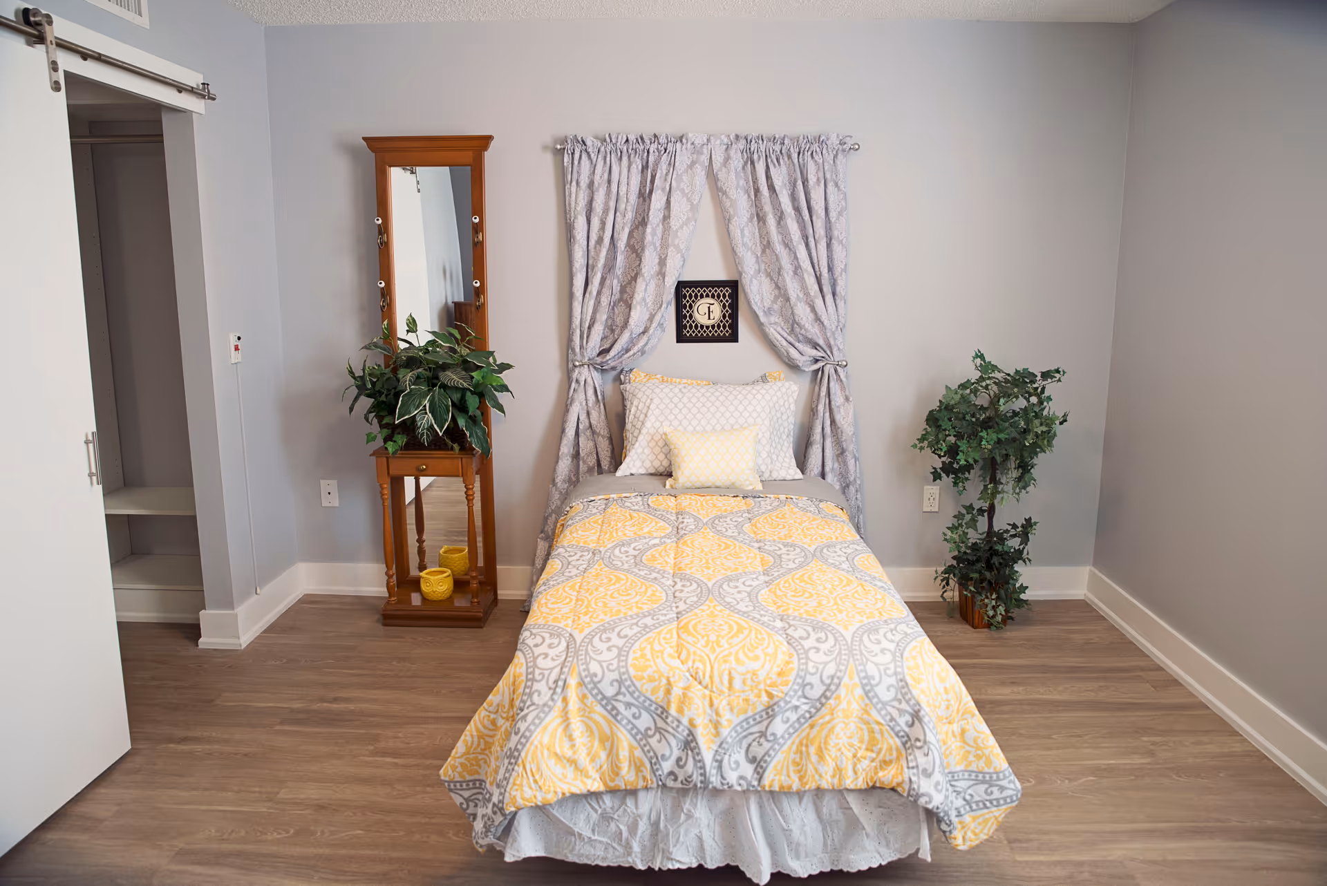 A single bed with a yellow and gray patterned comforter and pillows is centered against a light gray wall. Behind the bed, there are gray patterned curtains tied back on either side and a small framed decorative piece on the wall. To the left of the bed is a wooden stand with a tall mirror and a green potted plant on top, along with two yellow candles on the bottom shelf. To the right of the bed is a green leafy potted plant. The room has light wood flooring and a sliding closet door on the left side.
