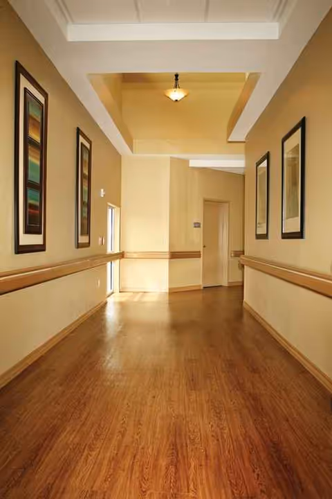 Long beige hallway with wooden floor, framed artwork, and handrails leading to a distant door.