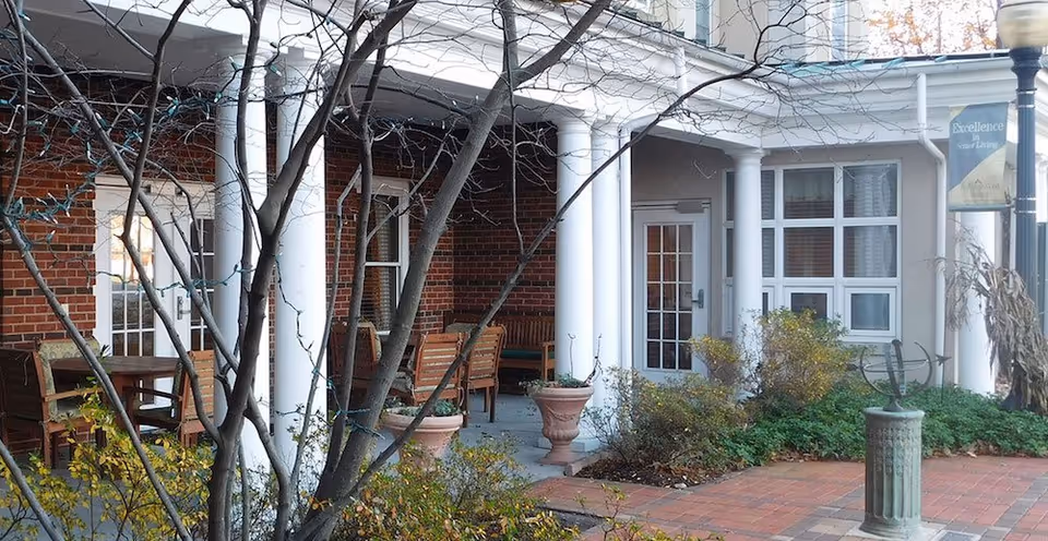 Outdoor covered patio area with white columns and brick walls, featuring wooden chairs and tables. Leafless tree branches and some green shrubs are visible in the foreground. A banner on a lamppost reads 'Excellence Senior Living.'