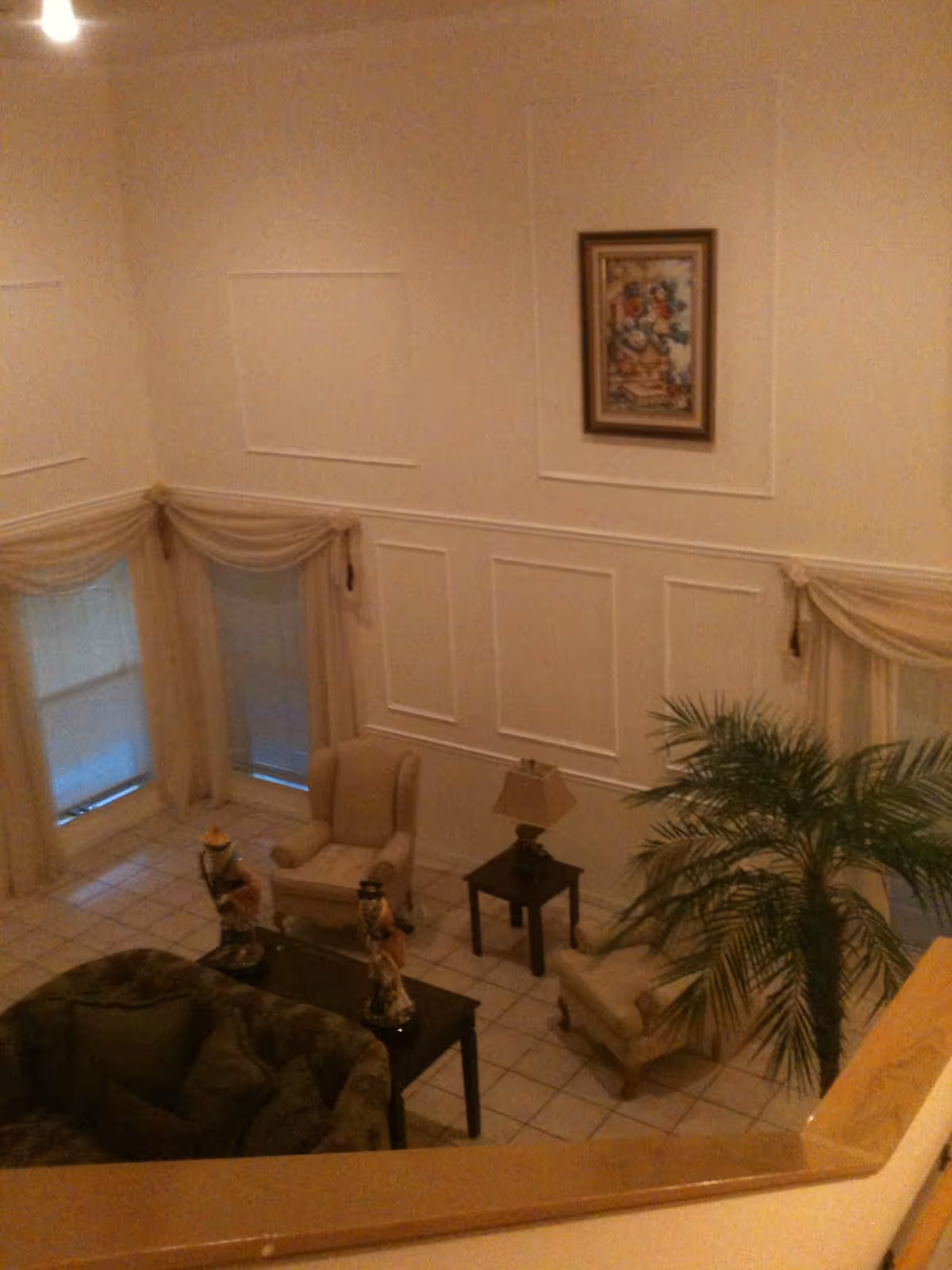 View down into a formal living room with armchairs, a sofa, side tables, a lamp, a potted palm, and framed art on paneled walls.